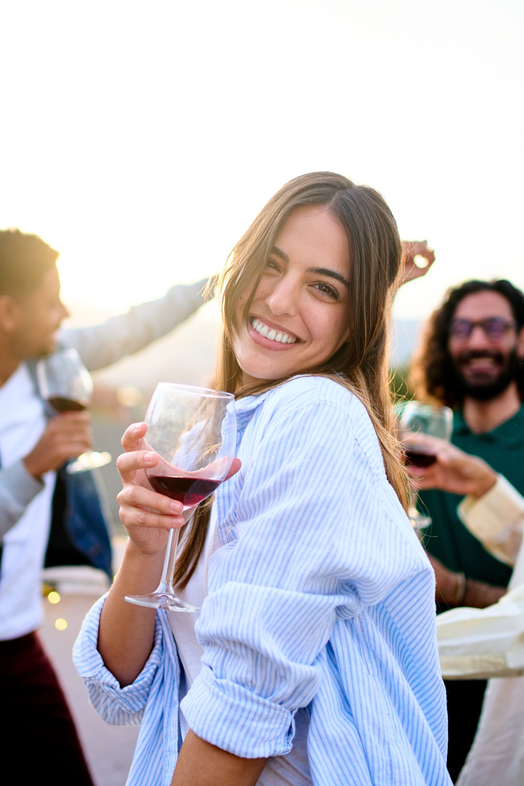 Woman smiling and holding a glass of wine at an outdoor gathering with friends in the background.